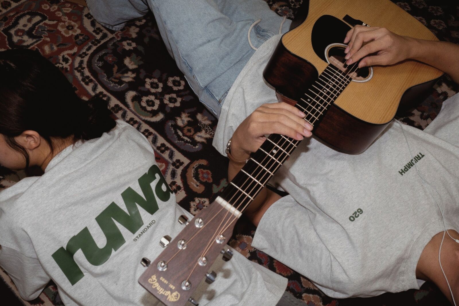 Two students on a rug with an acoustic guitar in Ruwa Standard and Triumph 620 tees